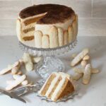 A three-layer tiramisu cake on a crystal cake stand with ladyfingers and a slice neatly arranged around it.