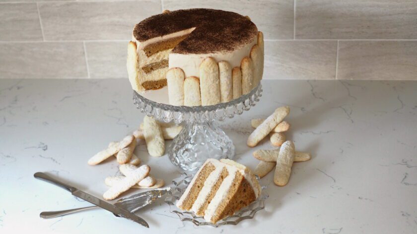 A three-layer tiramisu cake on a crystal cake stand with ladyfingers and a slice neatly arranged around it.