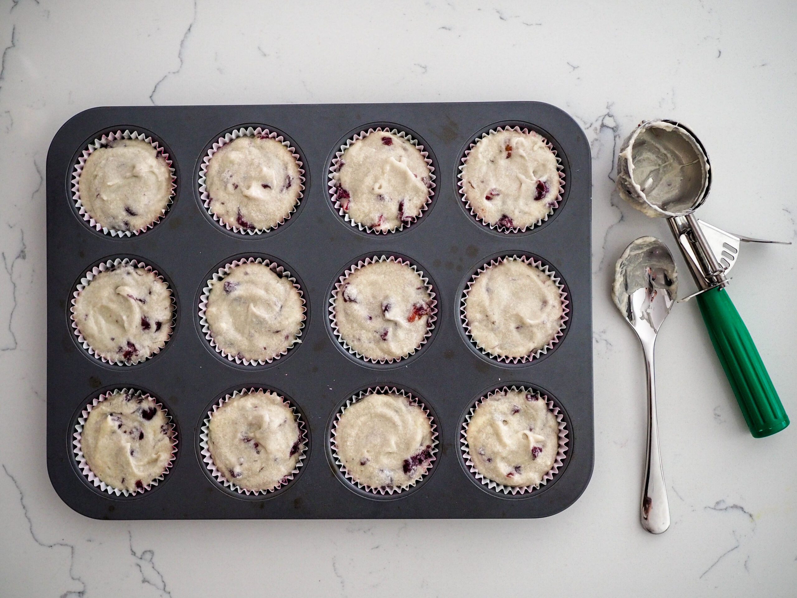 A muffin tin with cherry almond cupcake batter filled to the brim of each baking cup. A scooper and spoon lay on the counter next to it.
