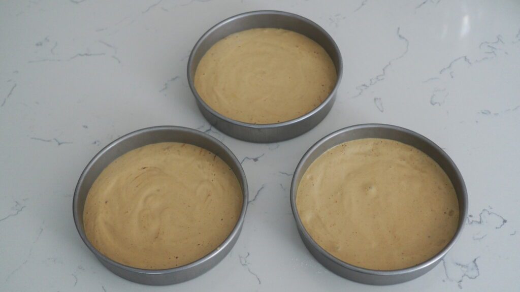 Three cake pans evenly filled with a light coffee sponge cake batter.
