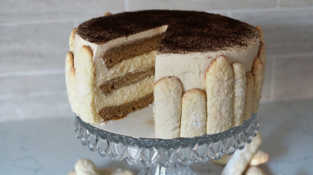 One large slice has been taken out from a tiramisu layer cake, decorated with ladyfingers of varying heights around the edges.