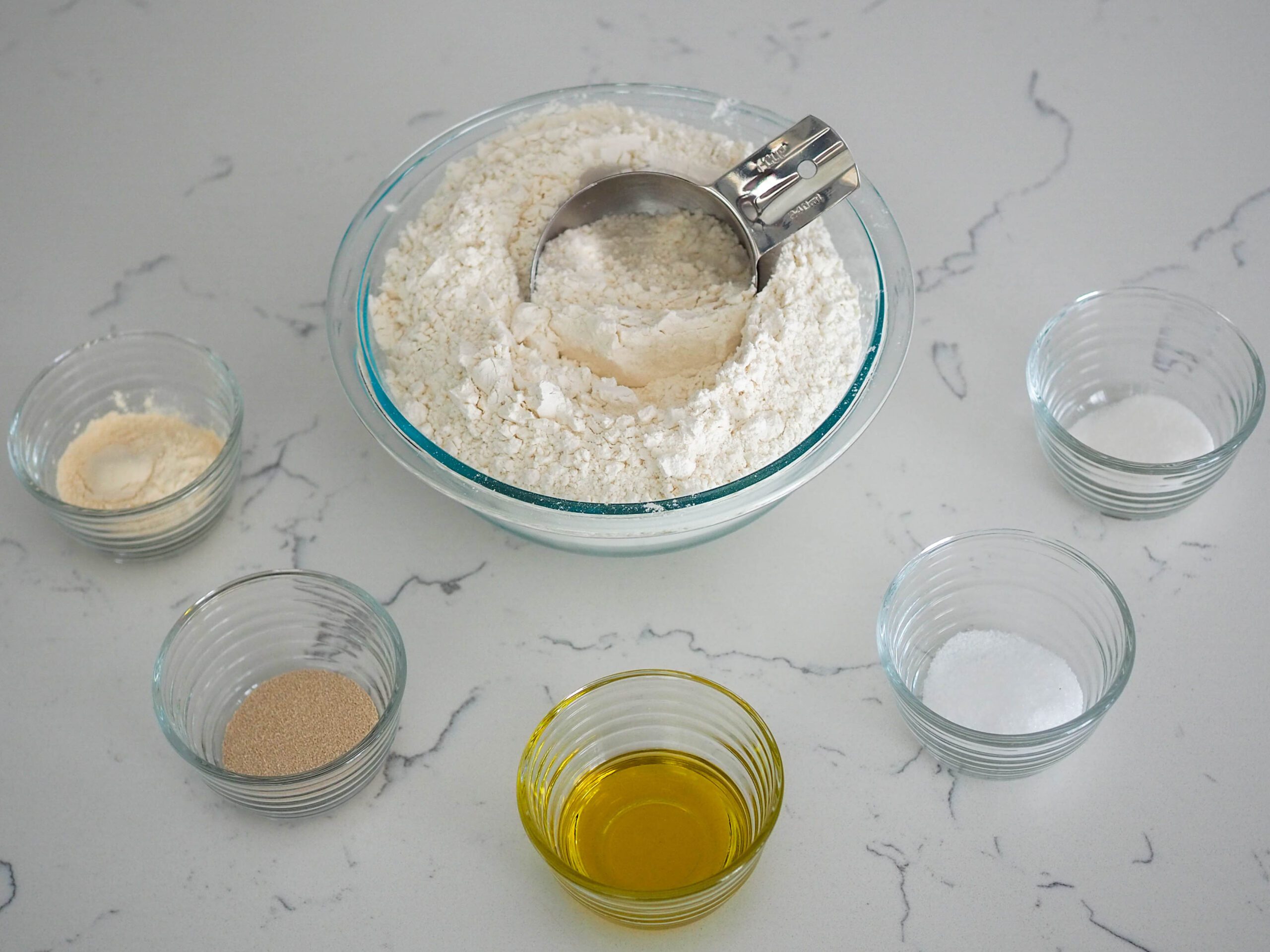 A bowl of flour with a measuring cup sticking out of it, along with small filled ingredient bowls surrounding it.