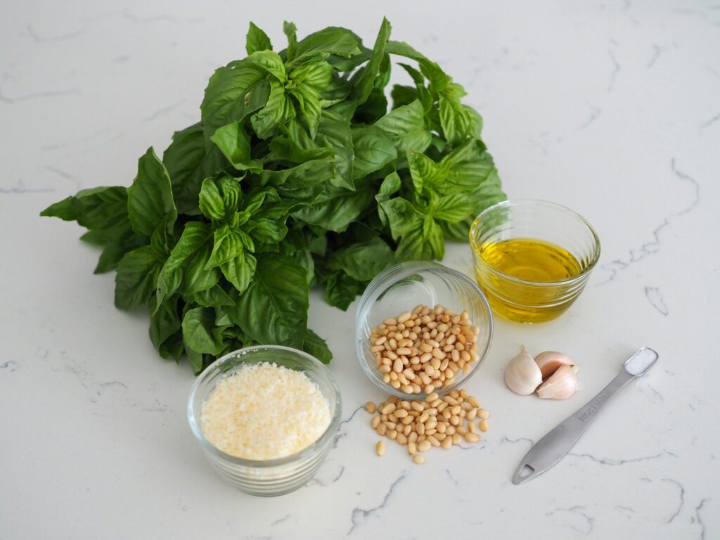 Basil, parmesan, pine nuts, olive oil, garlic, and salt on a white quartz counter.