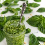 An open jar of pesto has a spoon sticking out of it, with basil leaves spread around the counter behind it.