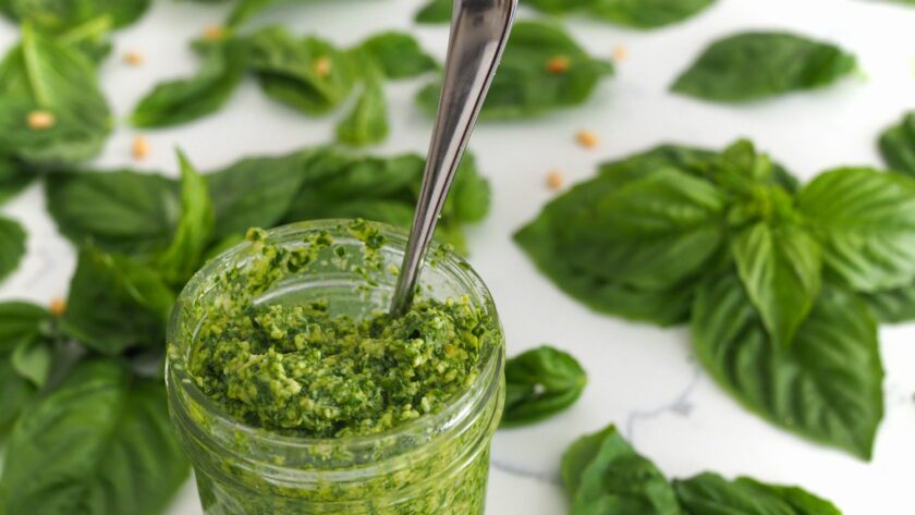 An open jar of pesto has a spoon sticking out of it, with basil leaves spread around the counter behind it.