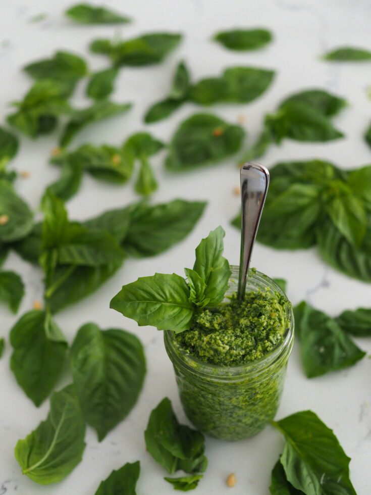 A jar of green pesto with a spoon coming out of it, surrounded by basil leaves.