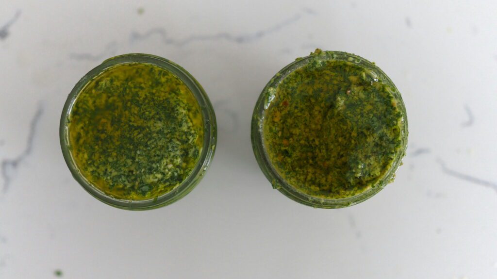 Two jars of basil pesto: The one on the left used raw pine nuts, and the one on the right used toasted pine nuts. There are deeper brown and yellow specks in the jar on the right, attributing to greater flavor.