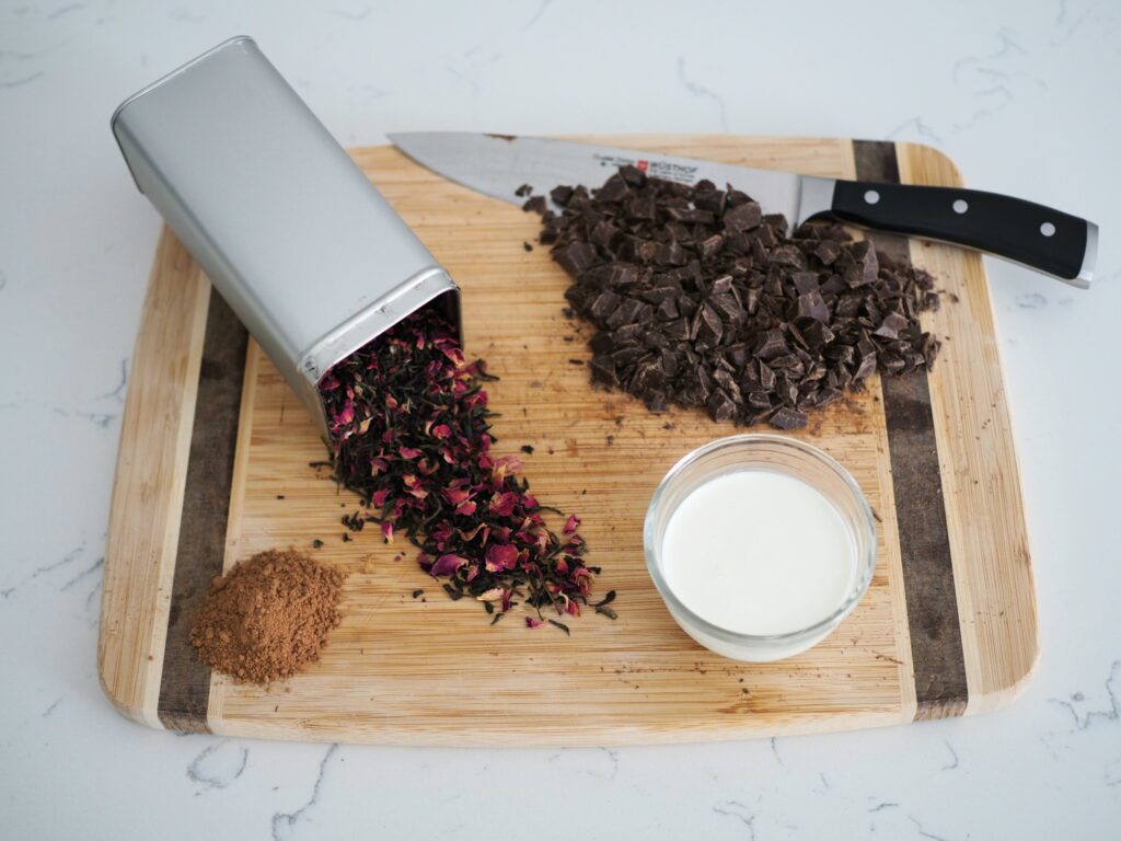 Chopped dark chocolate, heavy cream, cocoa powder, and rosy earl grey tea on a wooden cutting board.