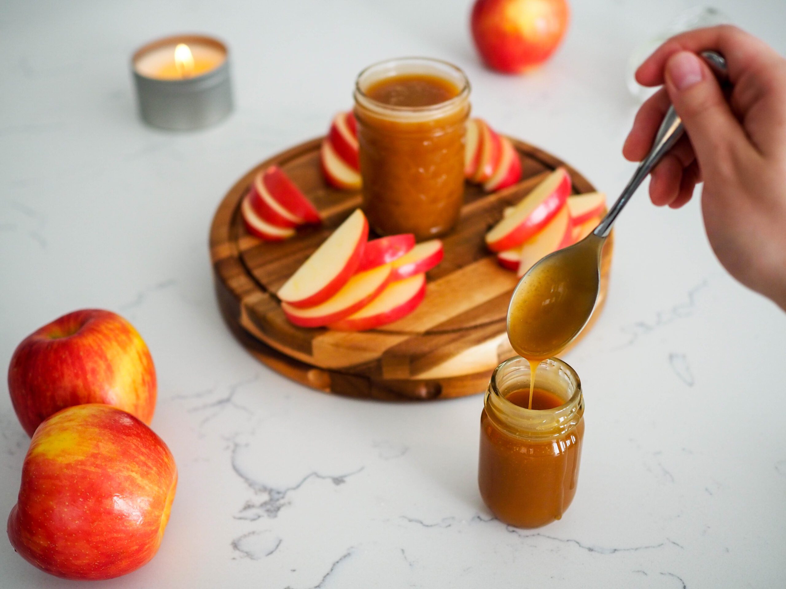 A hand dips a silver spoon into a jar of caramel, with sliced apples and another caramel jar behind it.