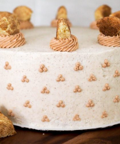 A full apple snickerdoodle cake, decorated in white cinnamon buttercream, with snickerdoodle cookies around the base of the wooden cake stand