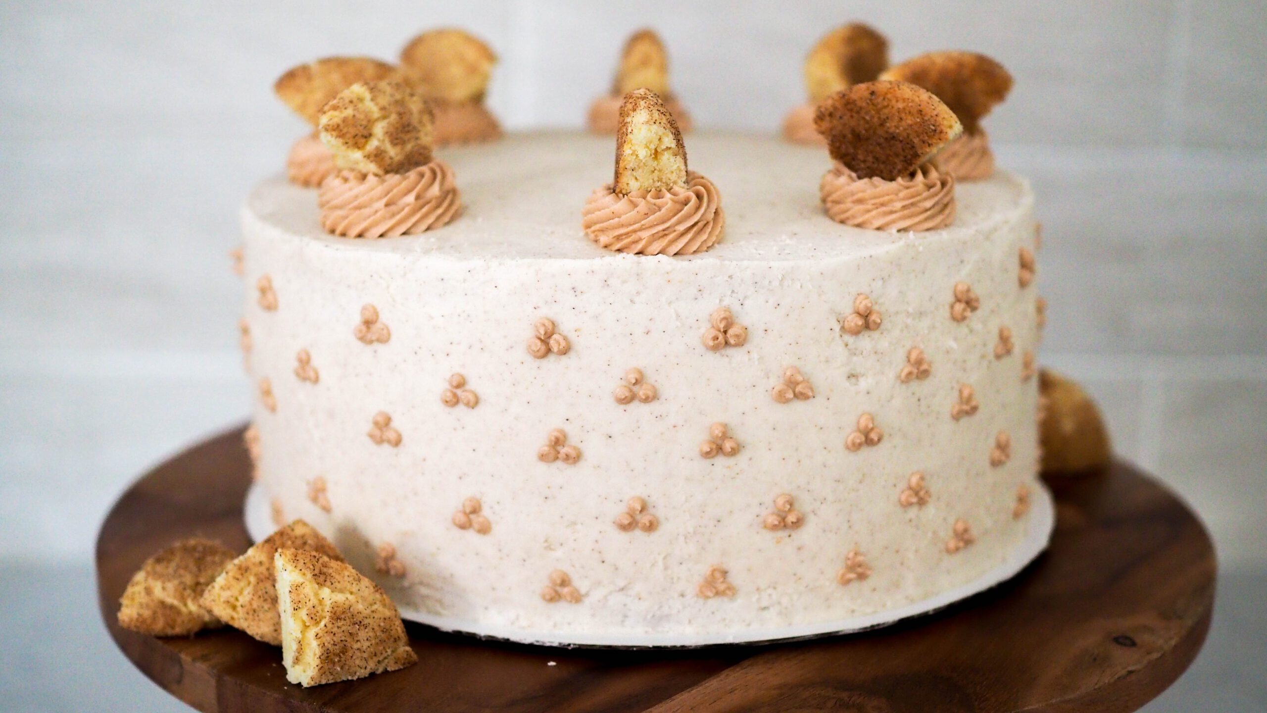 A full apple snickerdoodle cake, decorated in white cinnamon buttercream, with snickerdoodle cookies around the base of the wooden cake stand