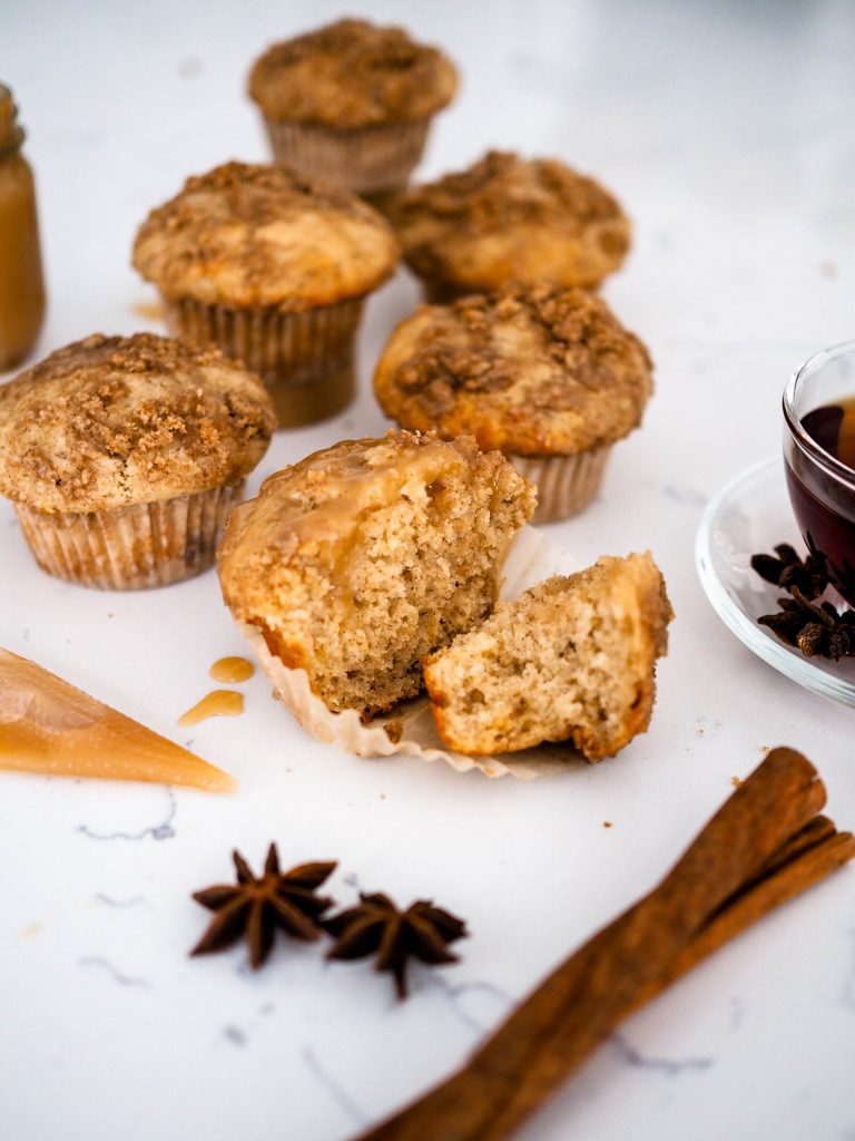 Five caramel chai muffins arranged on a white quartz counter with cinnamon sticks, star anise pods, caramel sauce, and tea surrounding them.