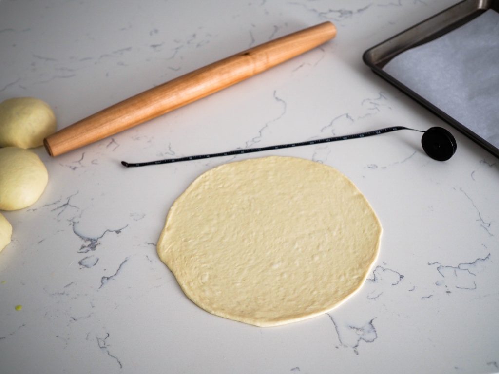 A rolled out ball of dough is on a white quartz counter, with a rolling pin, measuring tape, and baking sheet nearby.