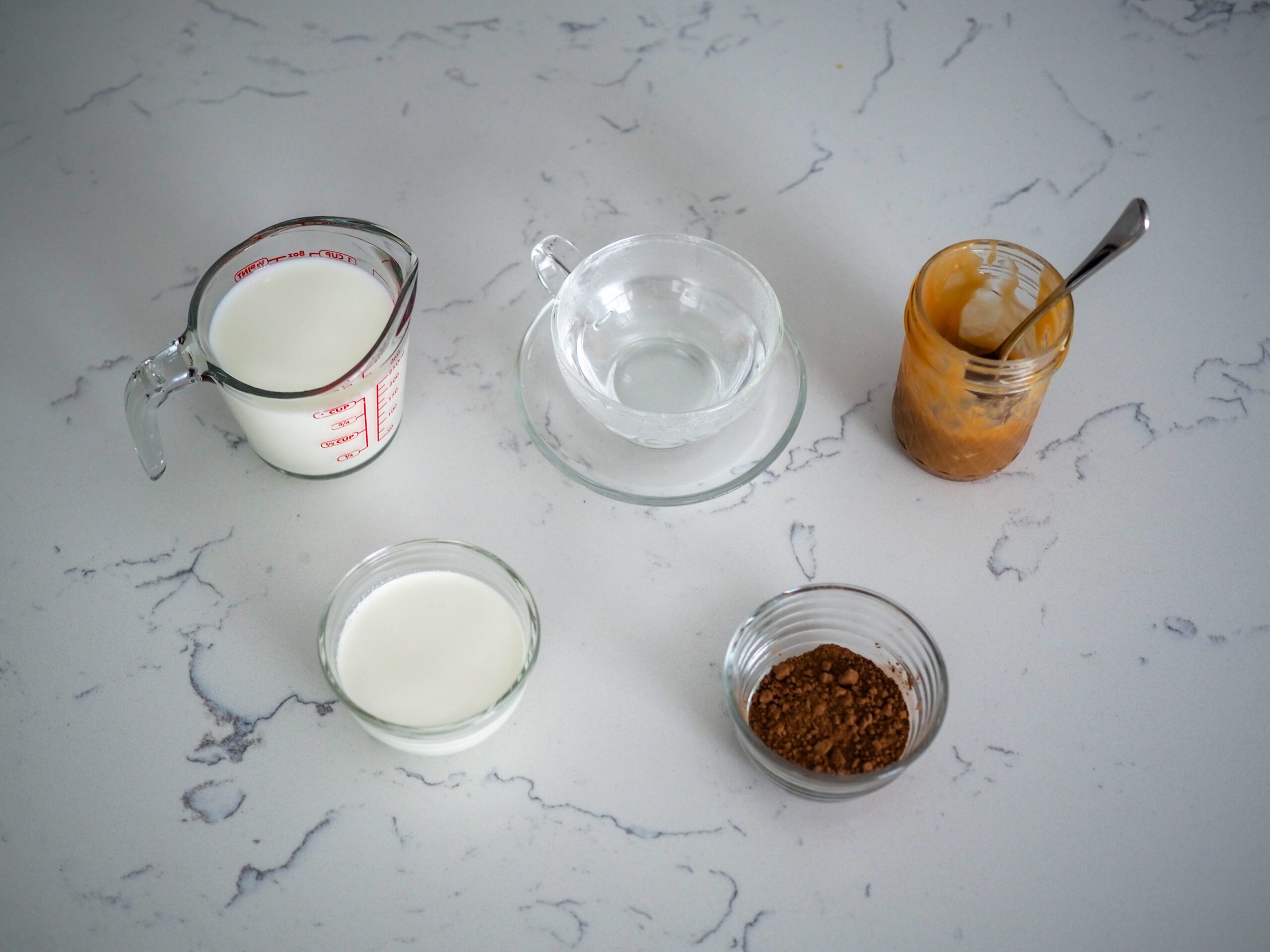 A measuring cup with milk, a tea cup with hot water, a jar of salted caramel, and two prep bowls filled with cocoa powder and heavy cream rest on a white quartz counter.