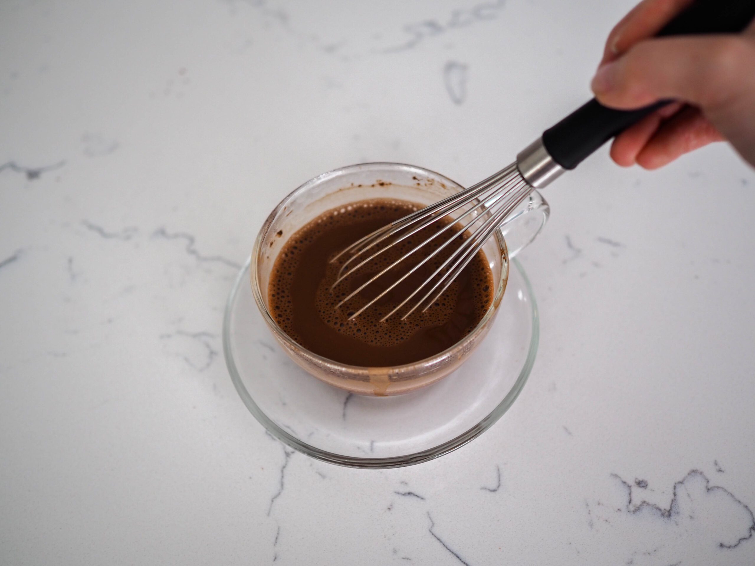 A whisk is resting in a clear tea cup filled with bloomed cocoa powder.