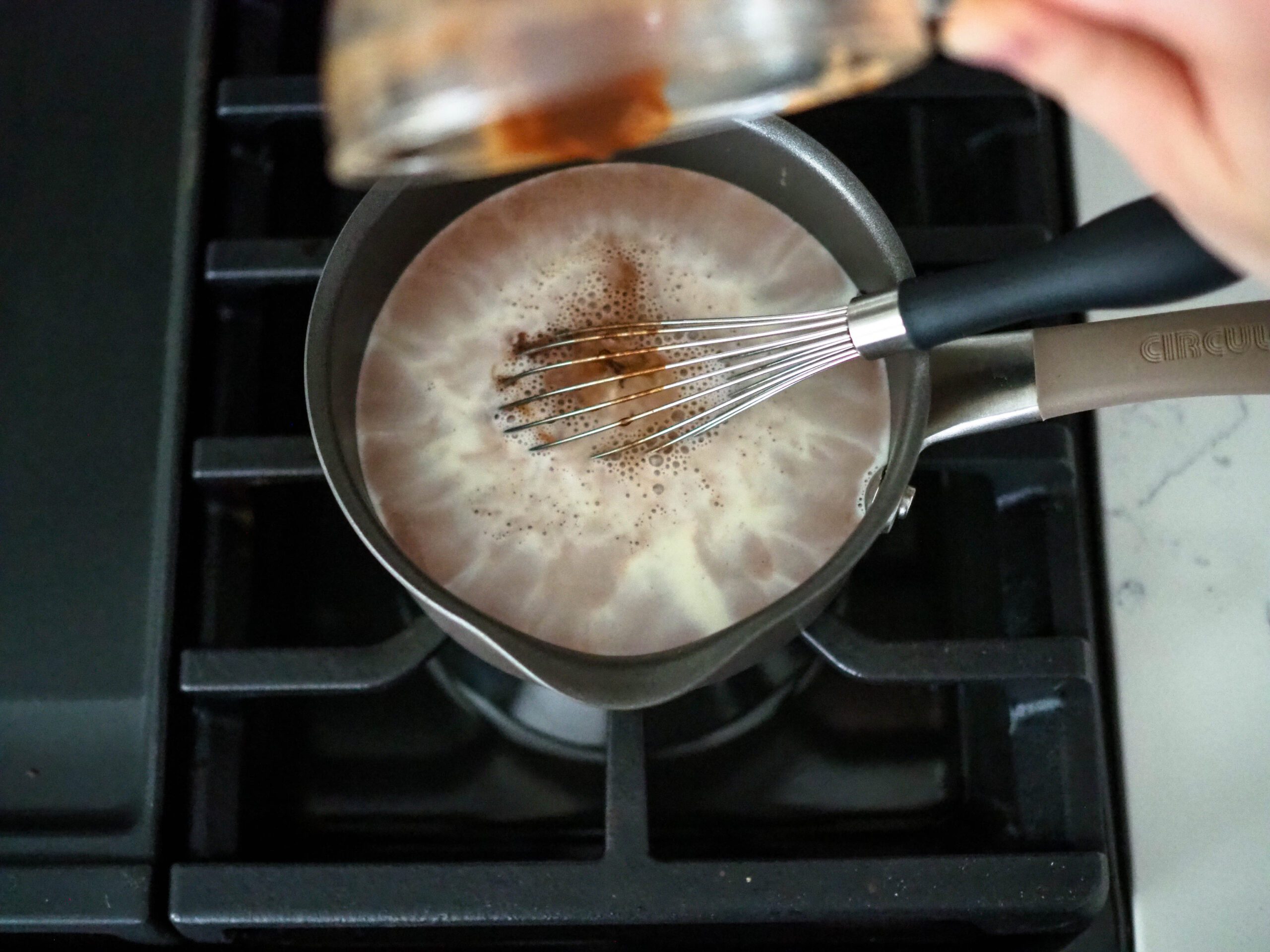 A hand pours bloomed cocoa powder into a small saucepan filled with cream and milk.