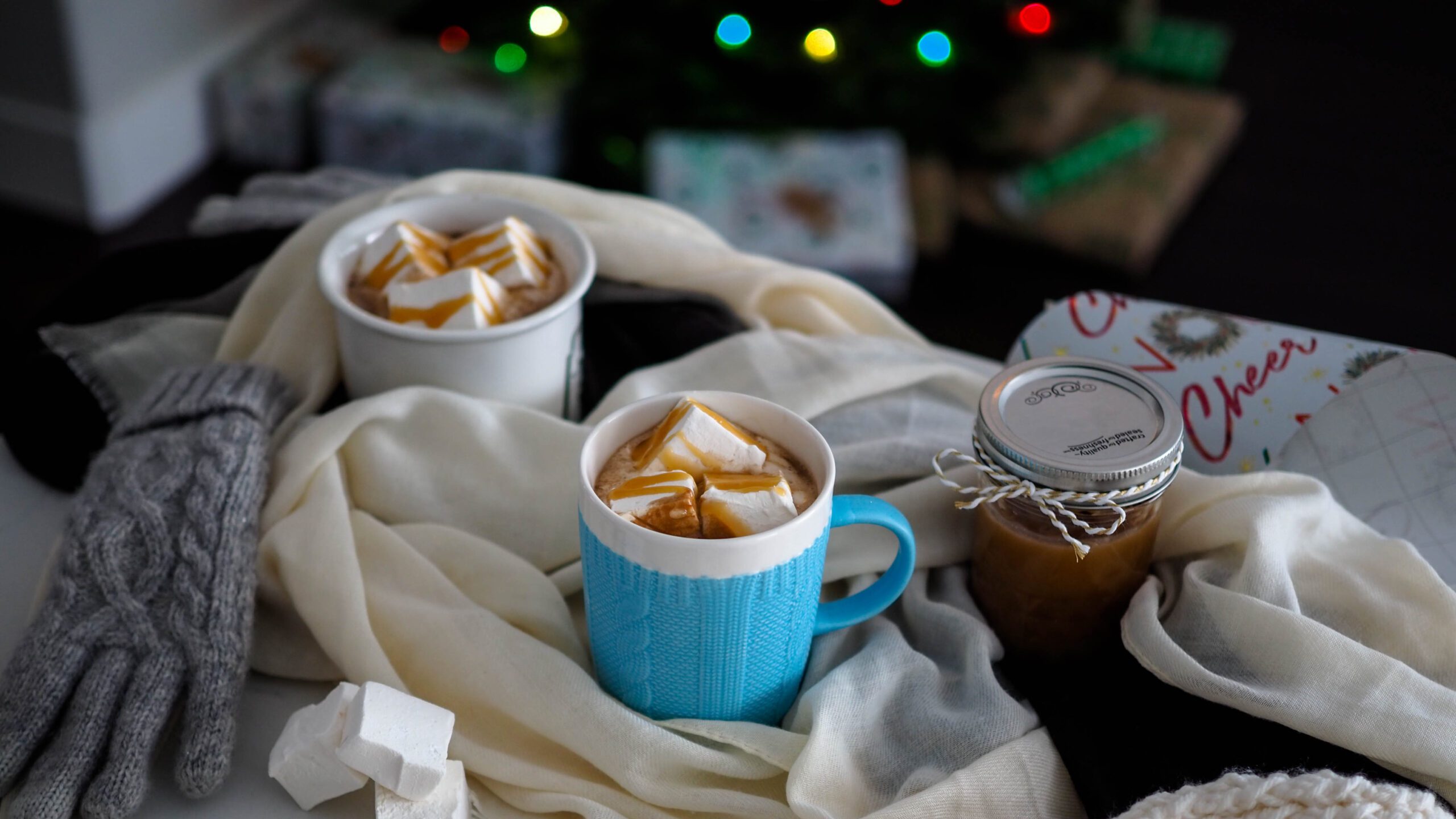 Two mugs of salted caramel hot chocolate on a busy kitchen counter filled with gloves, a scarf, and wrapping paper in front of a Christmas tree.