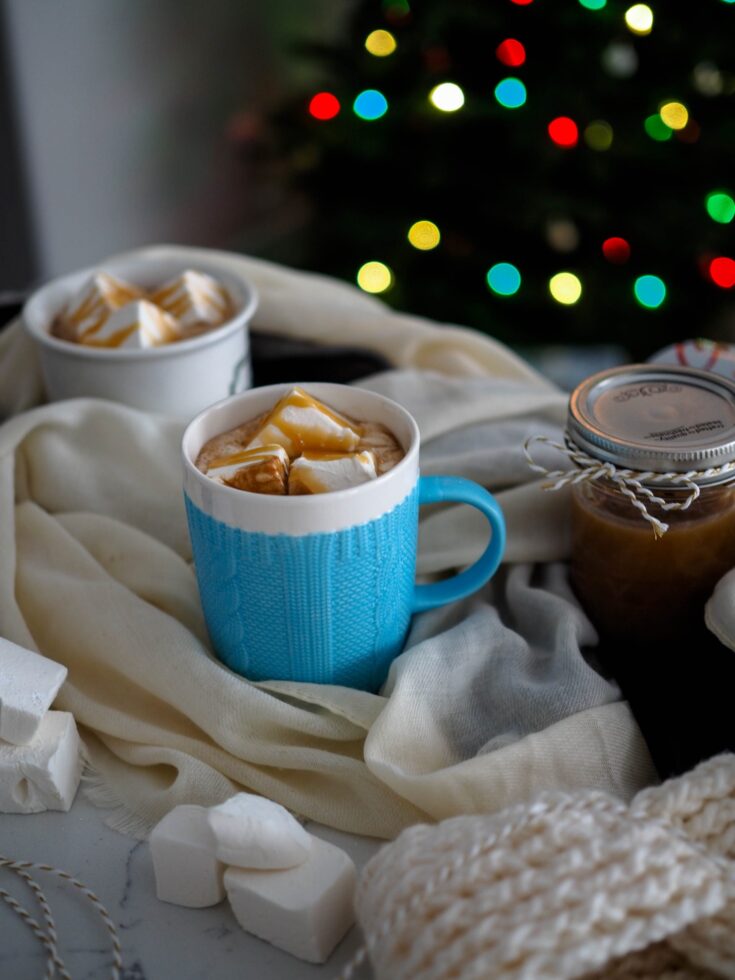 A textured blue mug is in the center of the frame, filled with hot chocolate and topped with marshmallows and caramel drizzles.