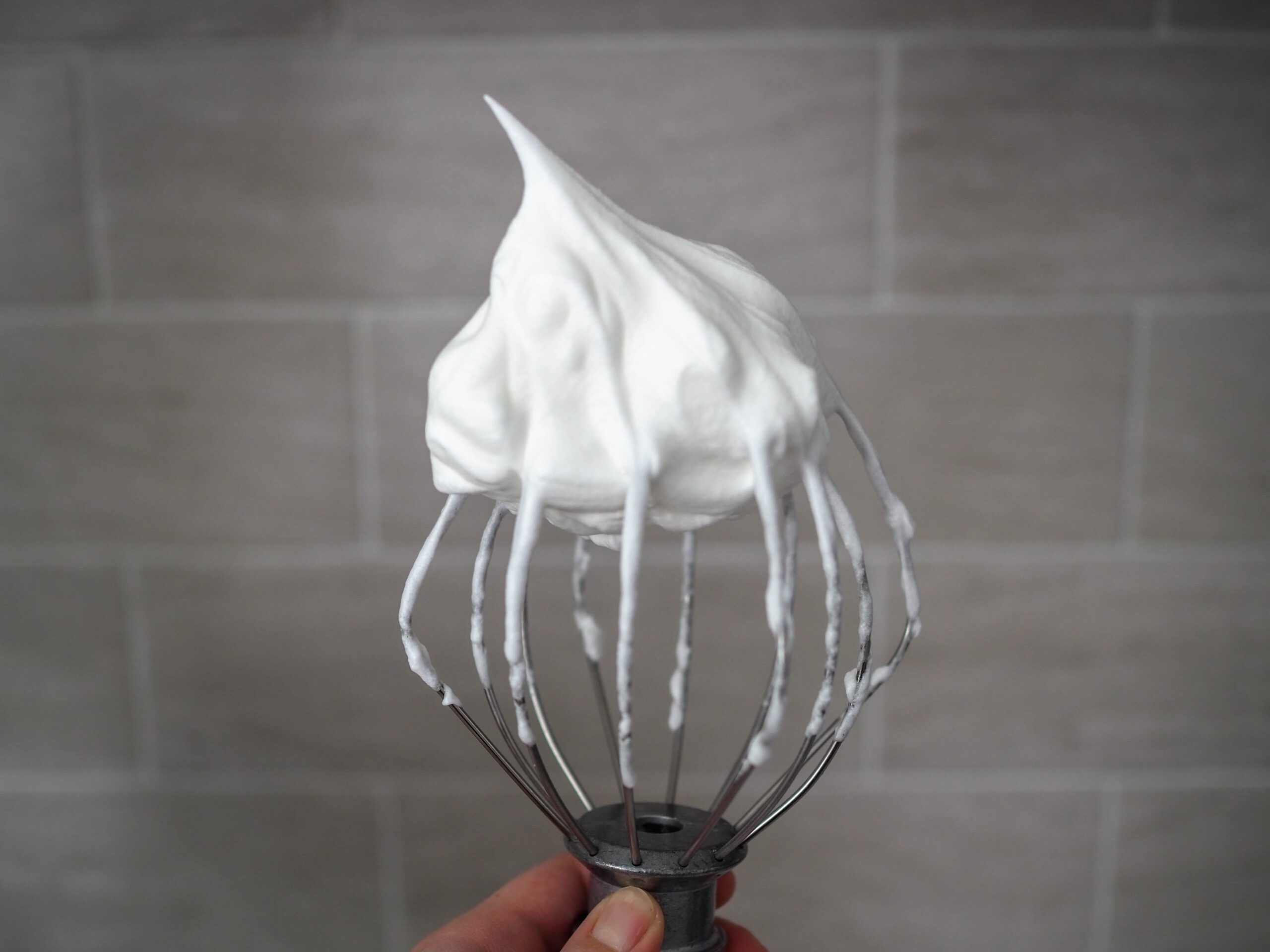 An upside down whisk attachment filled with meringue whipped to stiff peaks. The meringue has a sharp point, signifying it is properly whipped.