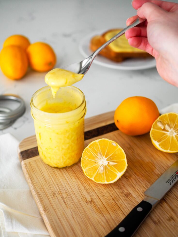 A hand lifts a teaspoon of lemon curd out of a full jar on a cutting board with cut lemons and a knife.