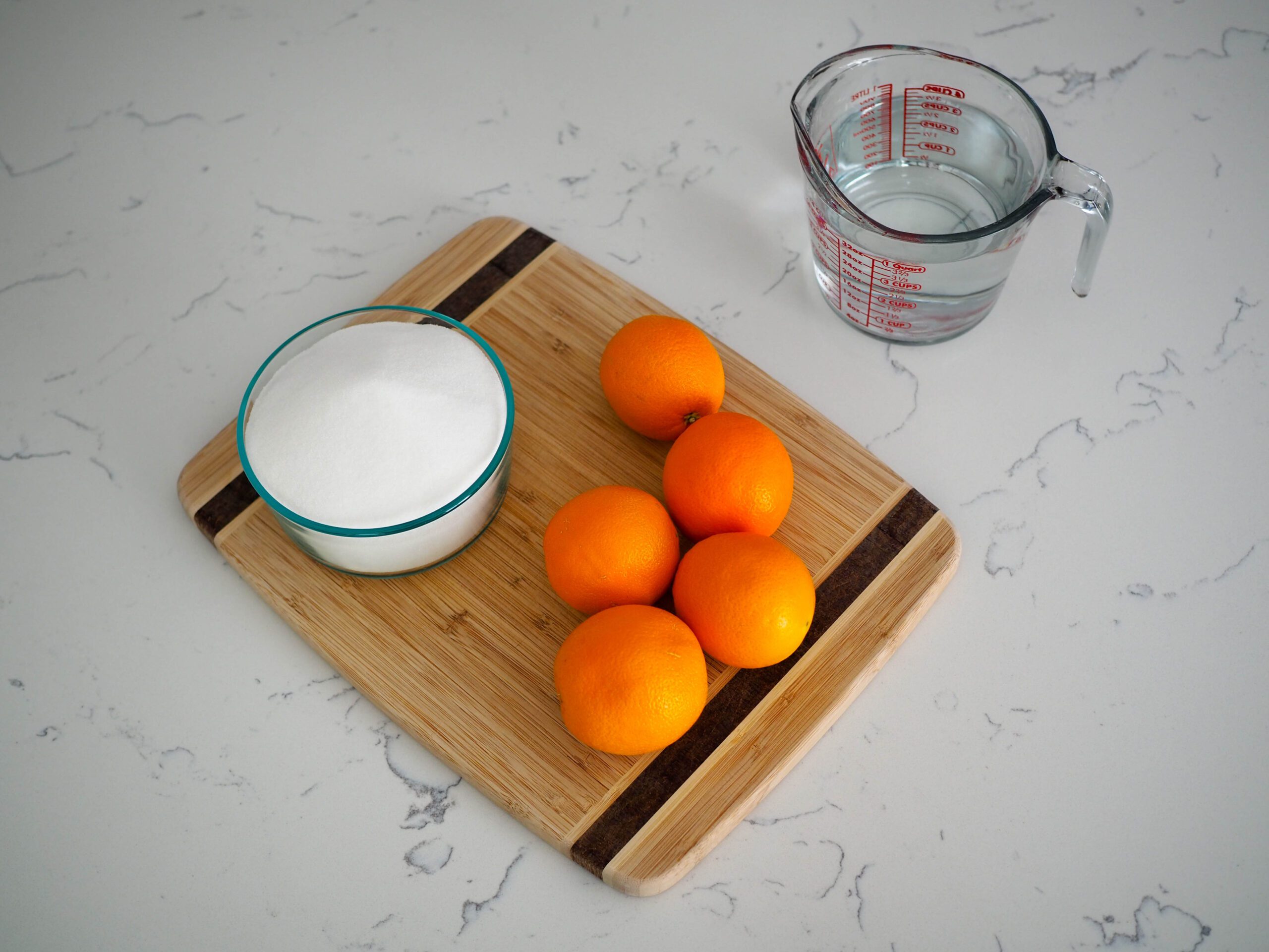 Five oranges, a bowl of sugar, and water on a quartz counter.