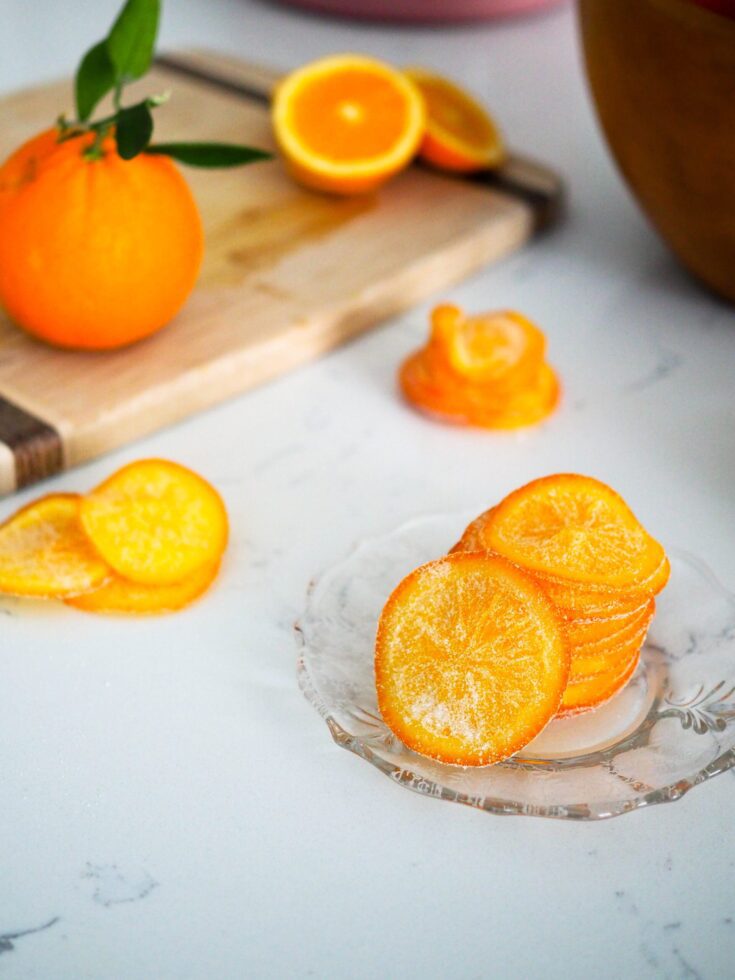 A stack of candied orange slices on a glass plate, with a bowl of oranges in the background.