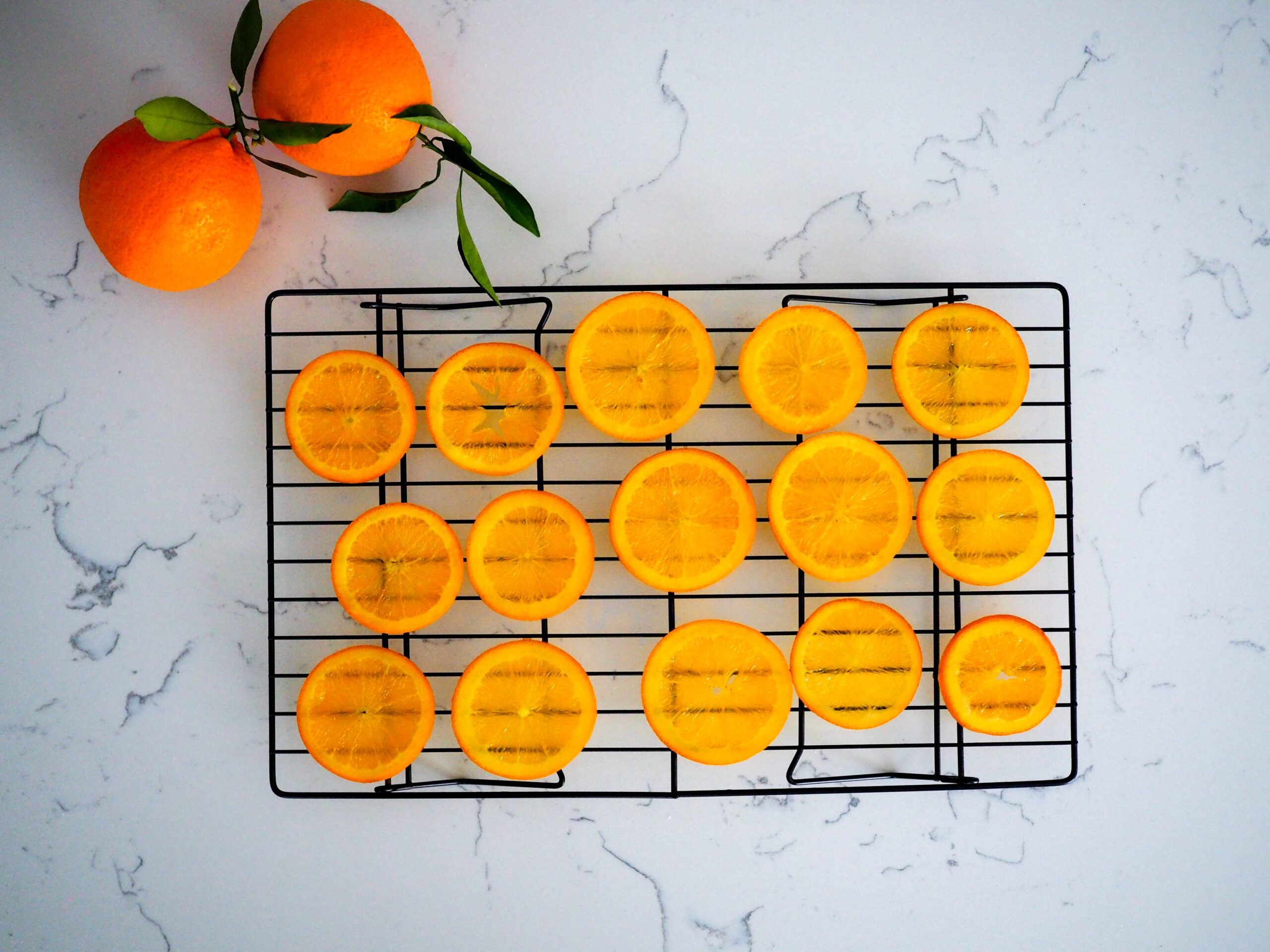 Fifteen orange slices dry on a wire rack with two leafy oranges beside them.