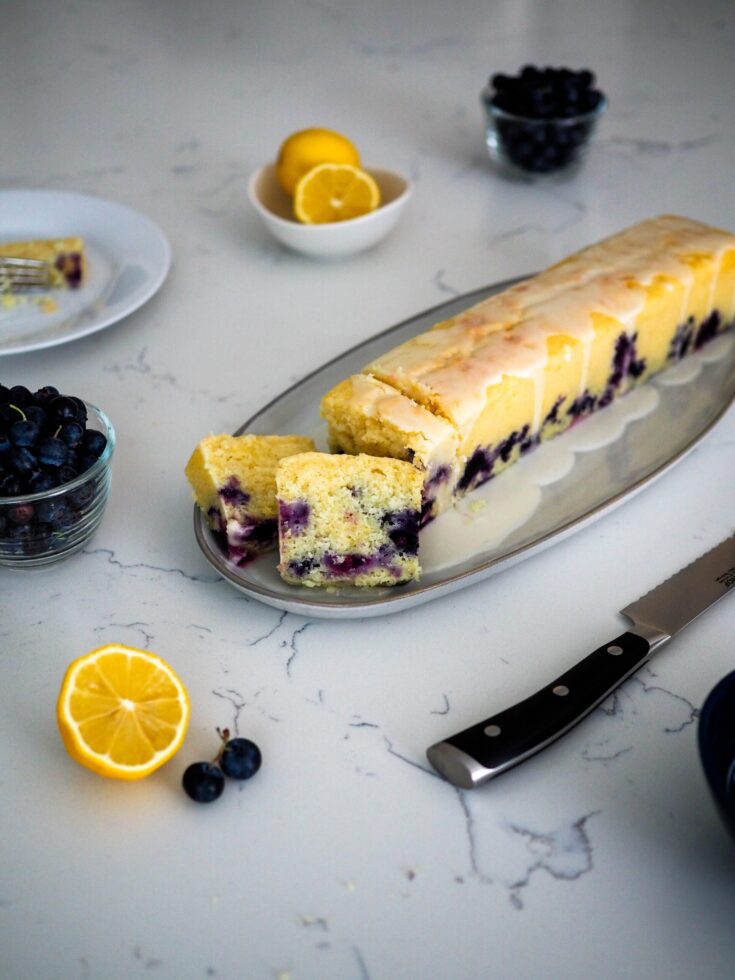 A cocktail-sized loaf of lemon blueberry loaf cake on a gray stoneware platter. A few slices are cut out of it, with a knife, blueberries, and lemons around it for staging.
