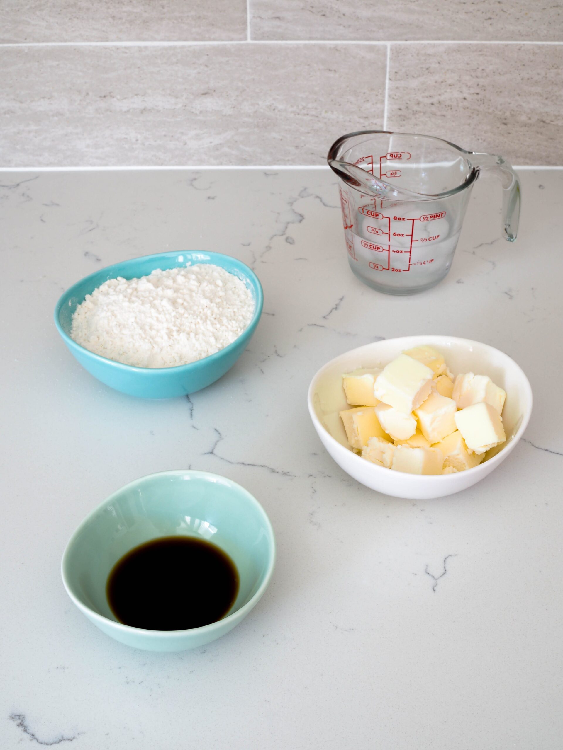 Flour, vanilla extract, butter, and ice cold water in small containers on a quartz countertop.