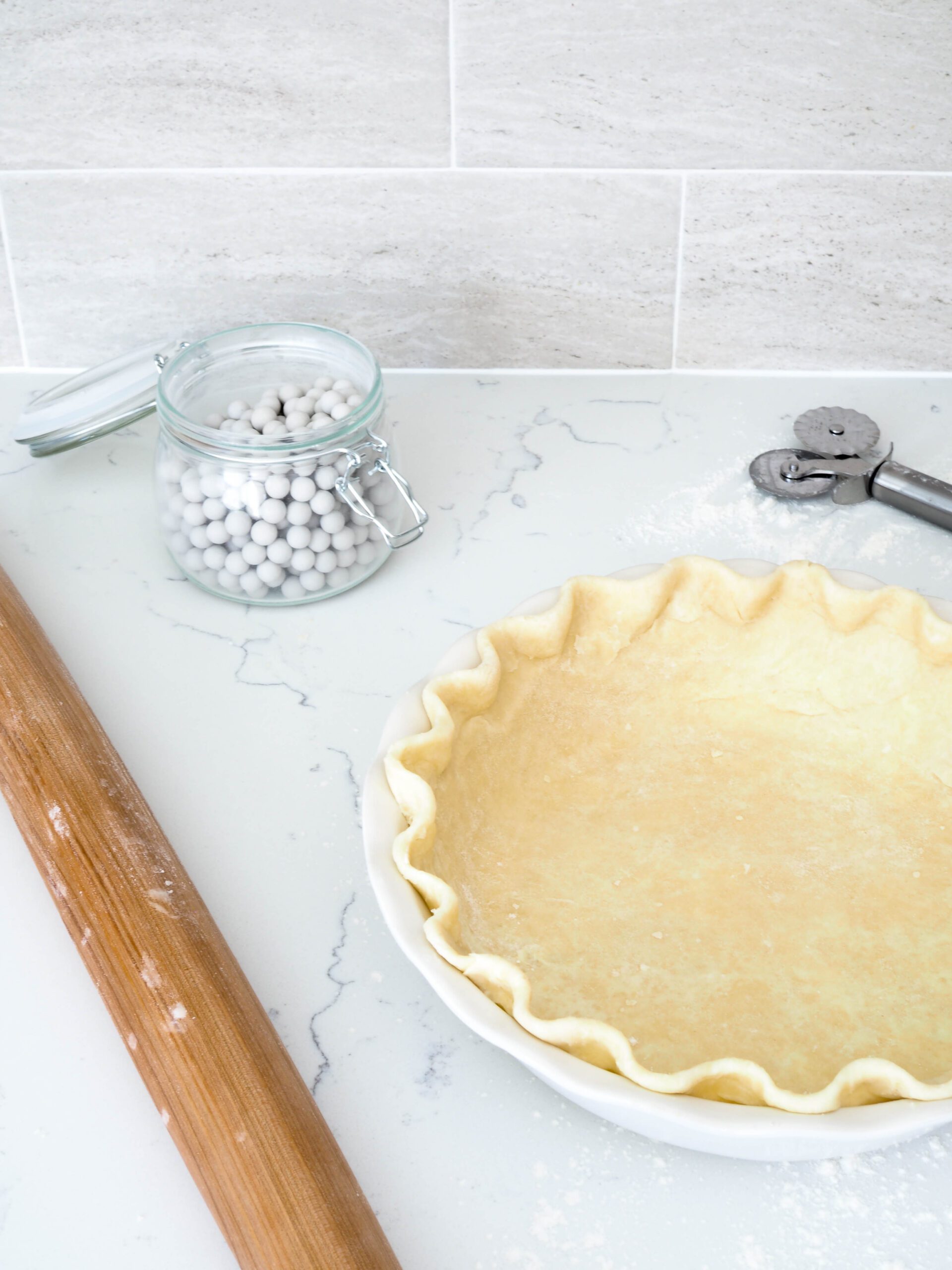 Two-thirds of a pie crust is in view in front of a gray backsplash with ceramic pie weights, a rolling pin, and a pie cutting tool.