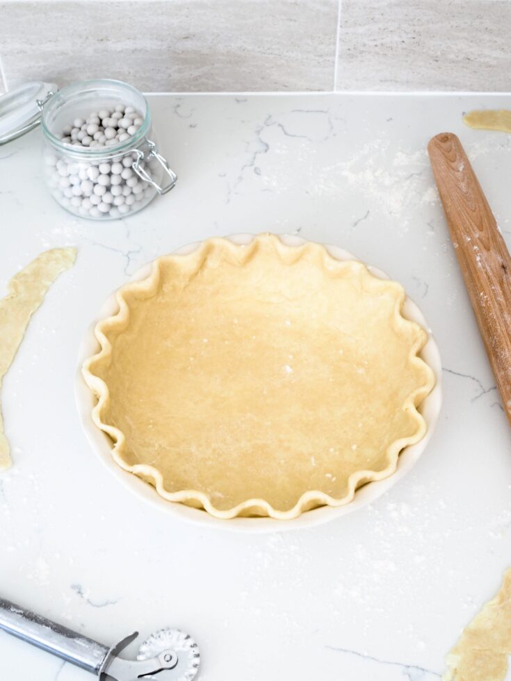 An unbaked pie crust with crimped edges on a white counter with a rolling pin, pastry cutter, and pie weights around it.