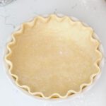 An unbaked pie crust with crimped edges on a white counter with a rolling pin, pastry cutter, and pie weights around it.