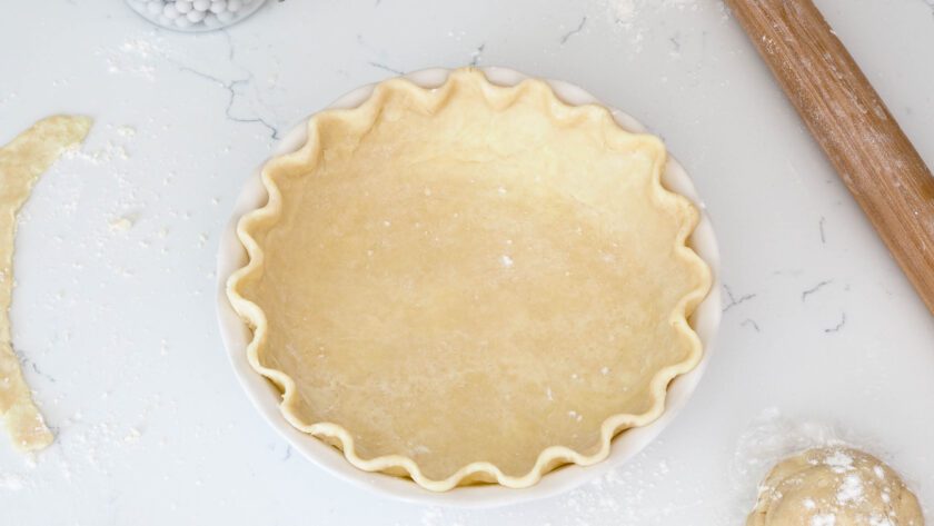 An unbaked pie crust with crimped edges on a white counter with a rolling pin, pastry cutter, and pie weights around it.