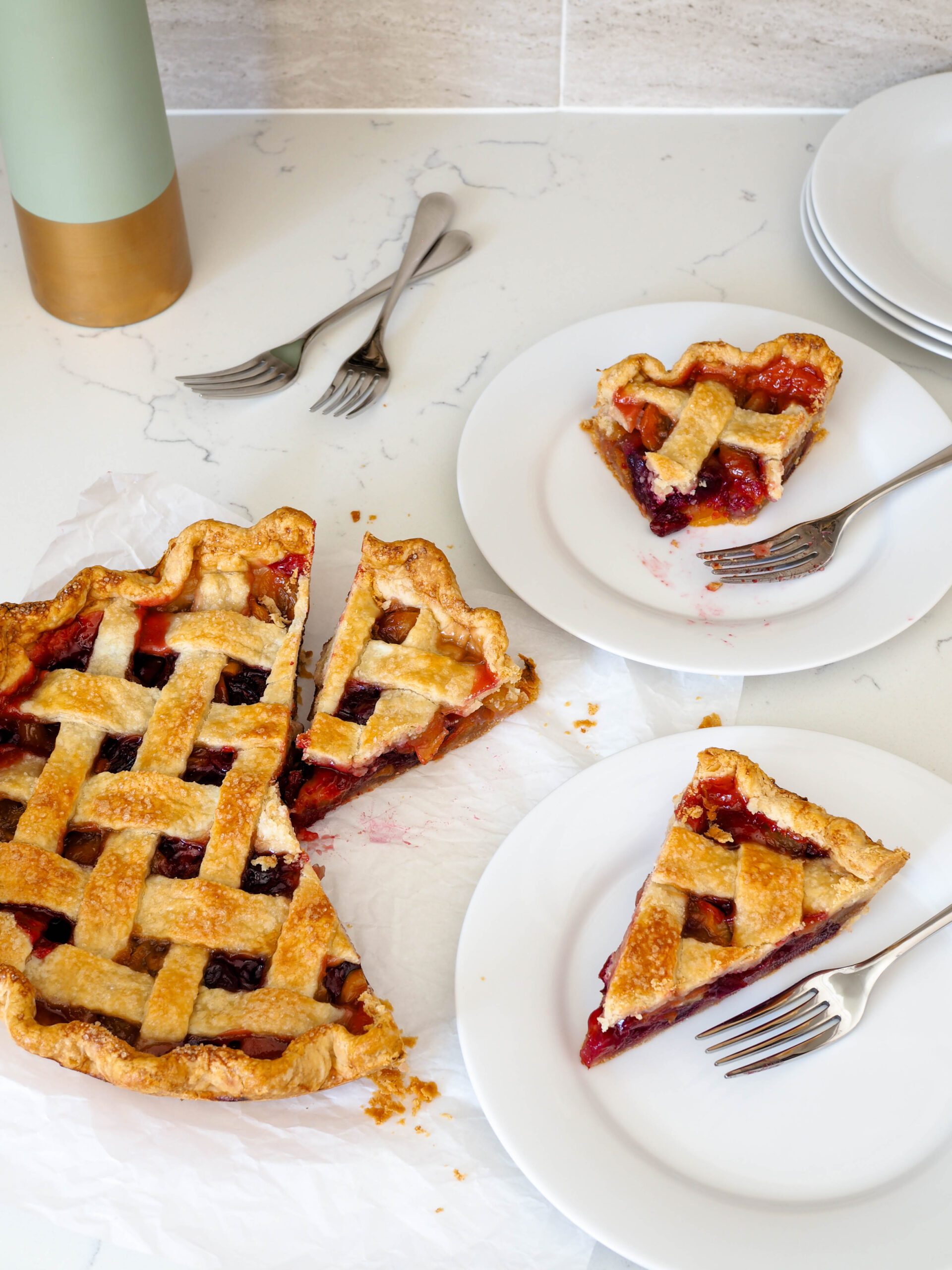 A cherry pie has three slices cut out of it, with two on plates with forks.