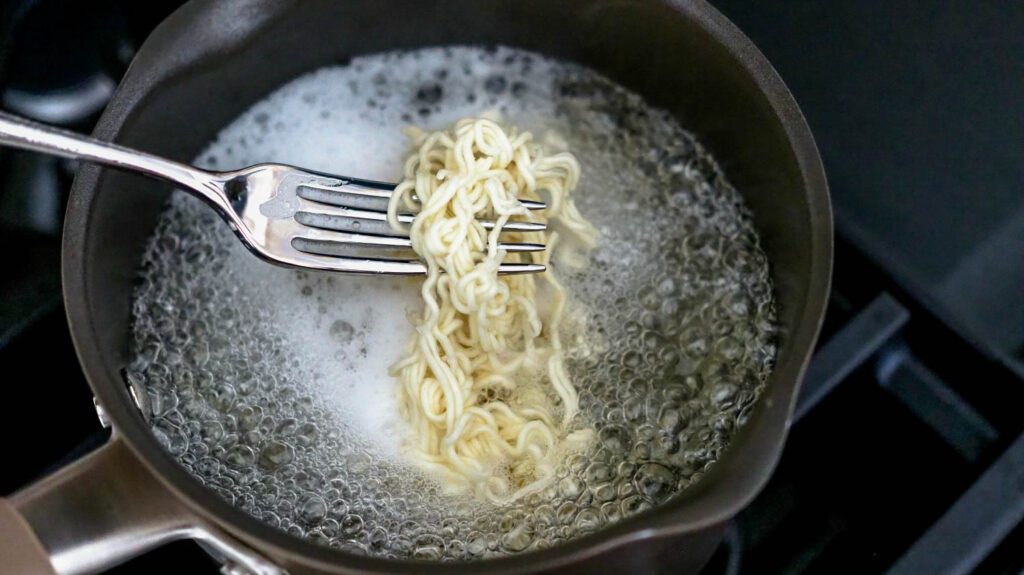 A fork holds up cooked ramen noodles above a pot of boiling water.