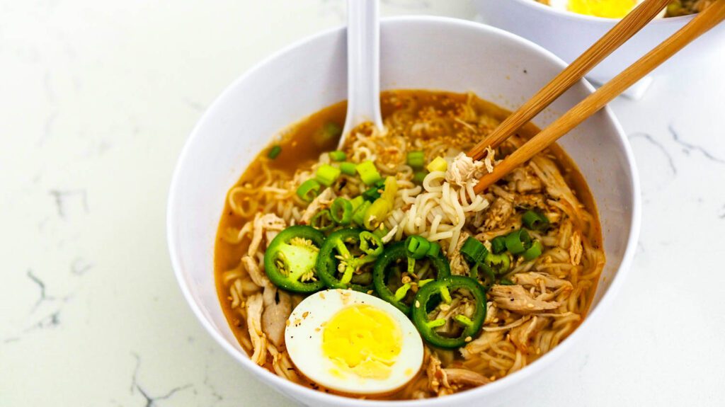 A pair of chopsticks is taking out a bunch of noodles and chicken from a bowl of ramen topped with jalapenos and an egg.