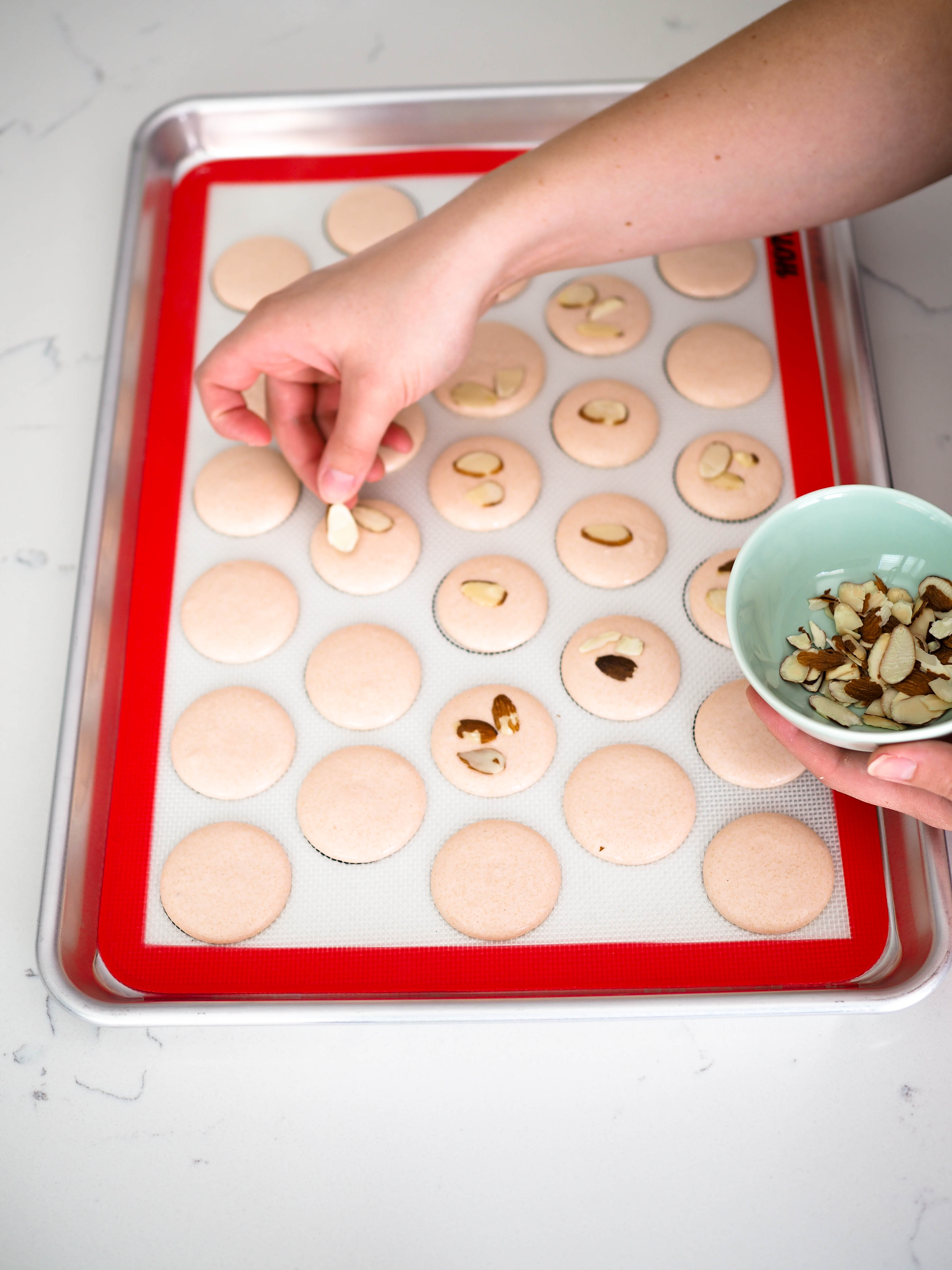 A hand places slices of almonds on top of piped macarons.