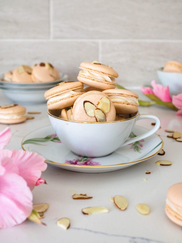 A pile of peach-colored macarons with slivered almonds in the shells inside of a dainty teacup with a pink flower blooming in the foreground.