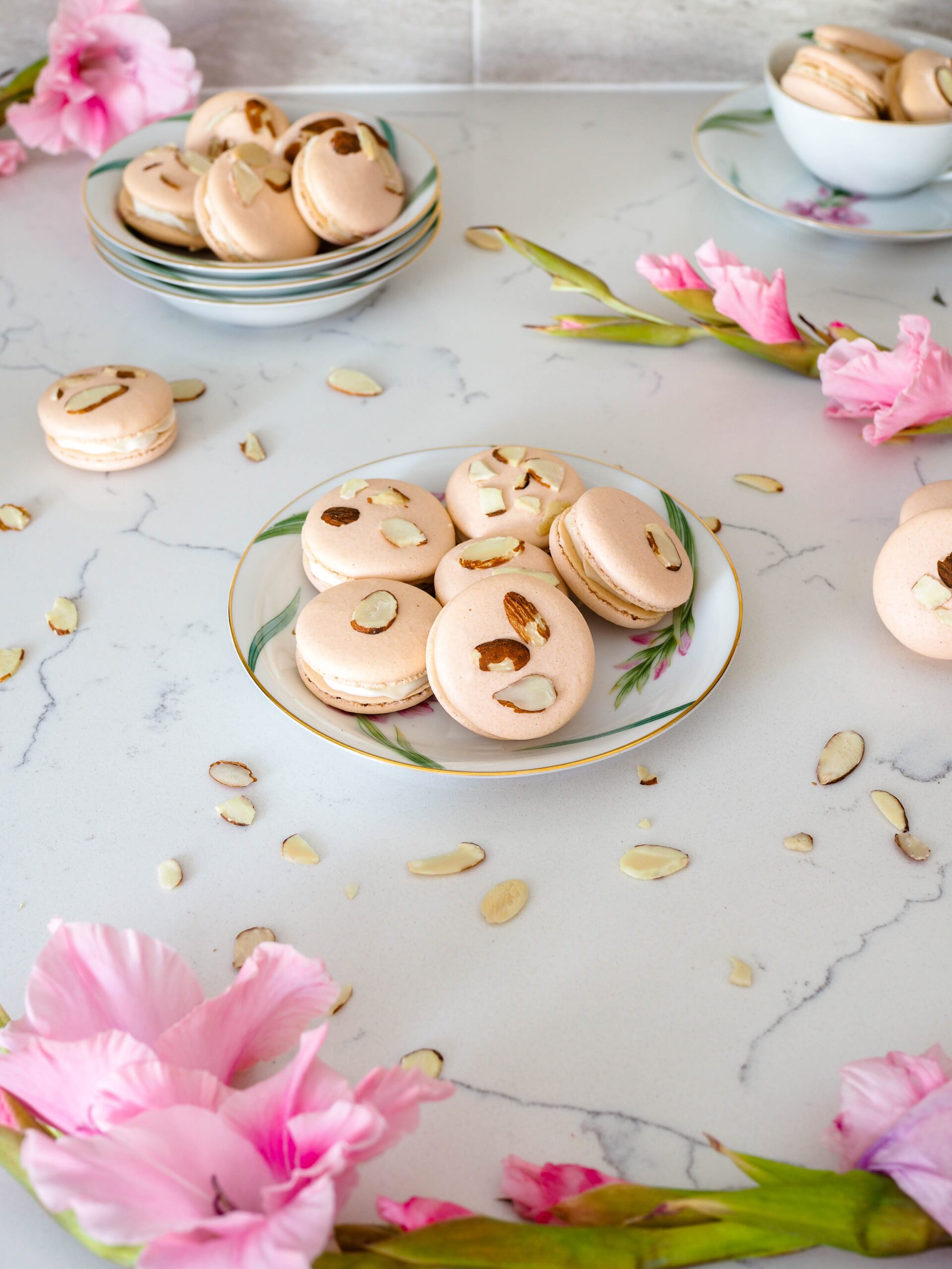 A handful of macarons is arranged on a china plate, with light pink gladiolus flowers and almond slivers surrounding them.