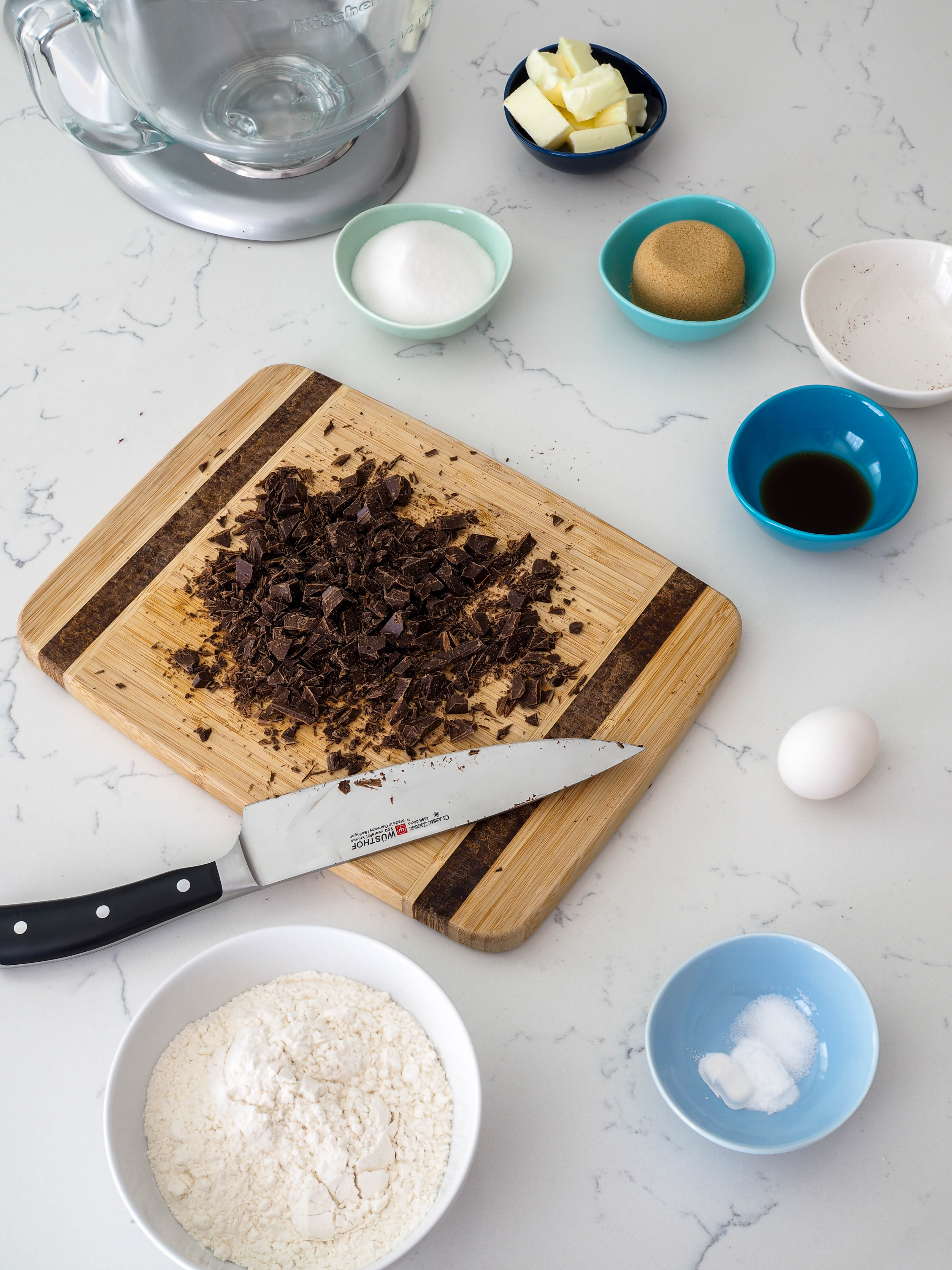A cutting board covered with roughly chopped chocolate and a Wusthof chef's knife is surrounded by cookie ingredients.