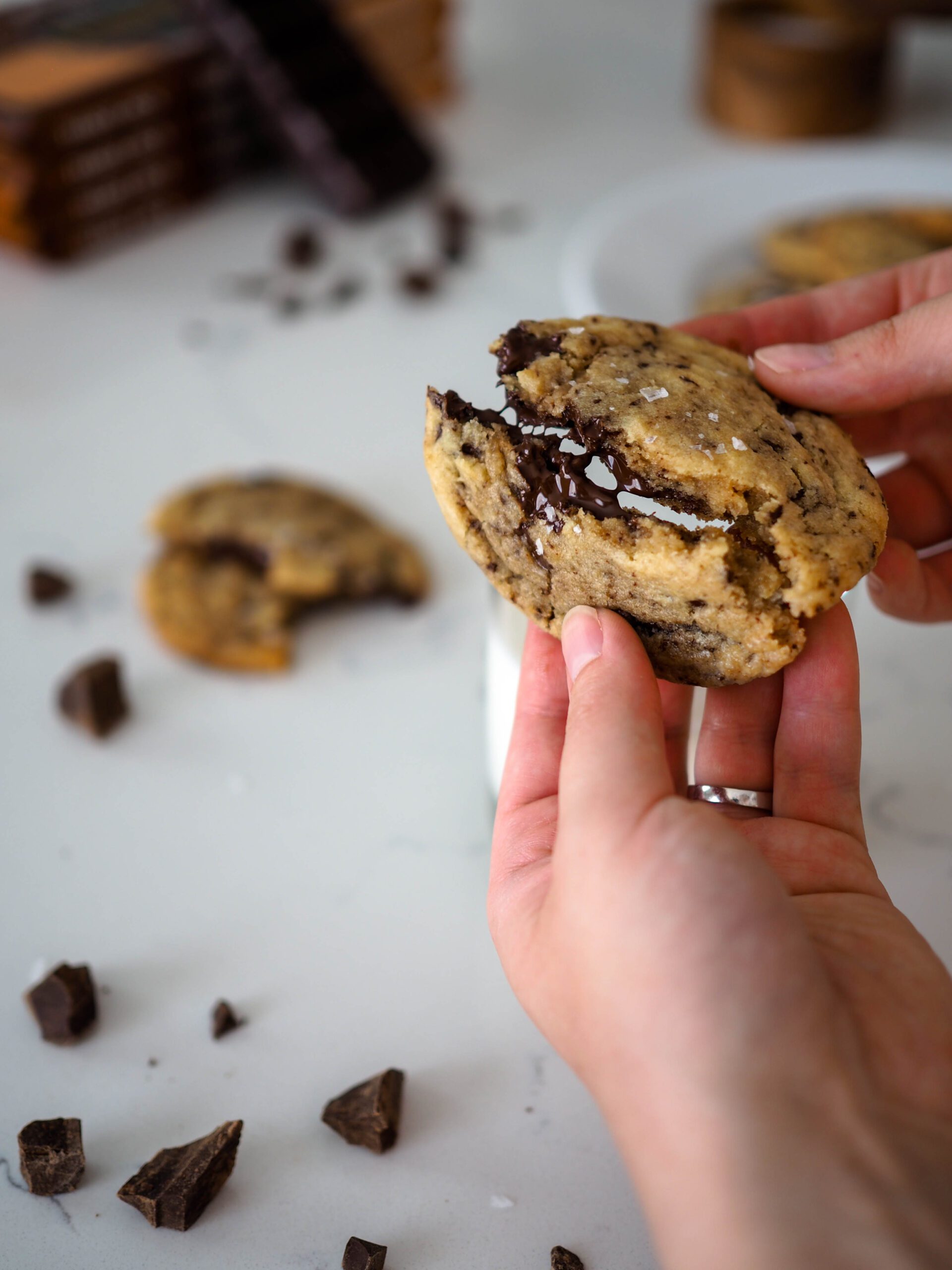 A dark chocolate chip cookie is broken in half by two hands.