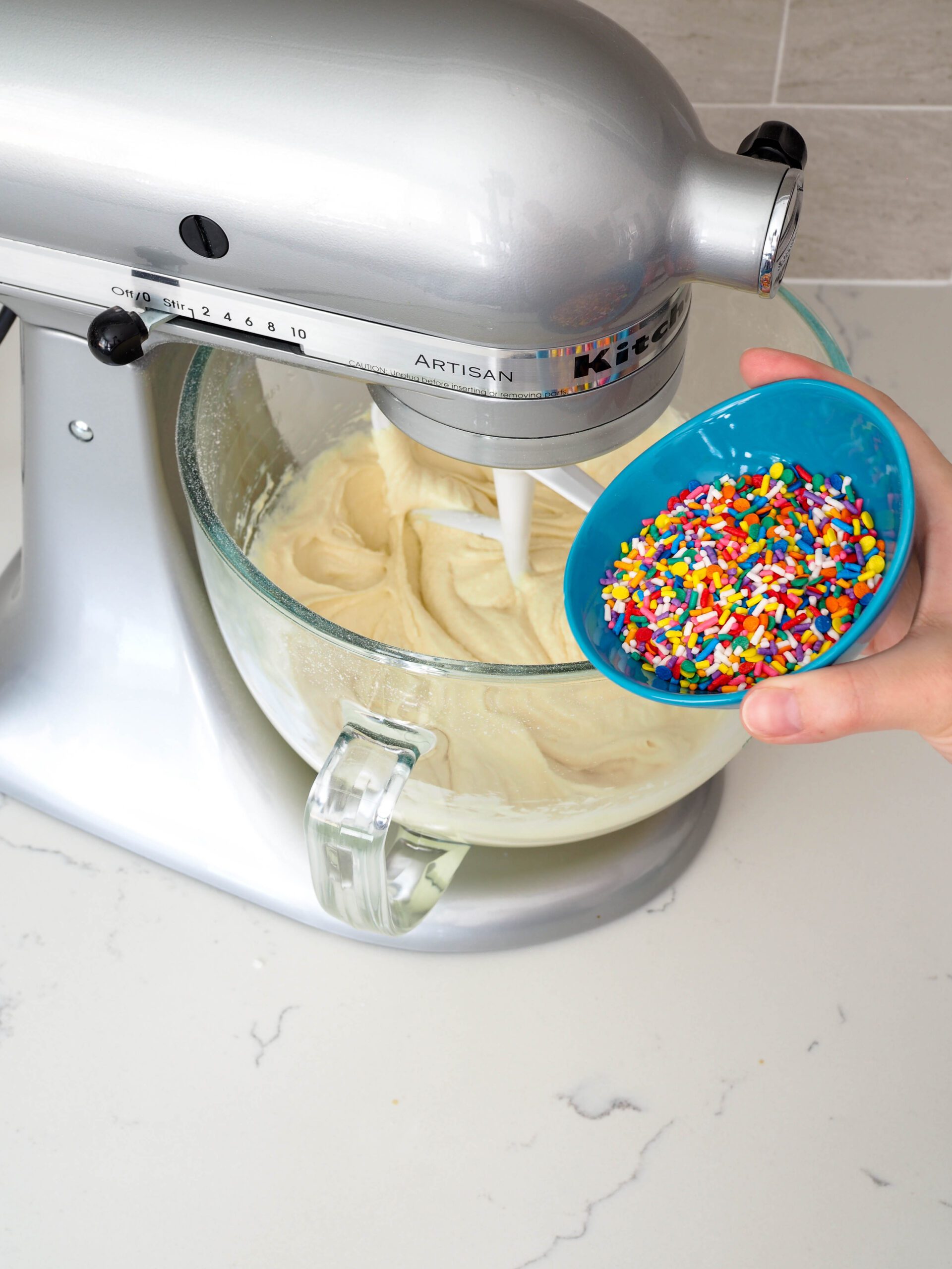 A bowl of sprinkles rests on the edge of a mixing bowl filled with cake batter.