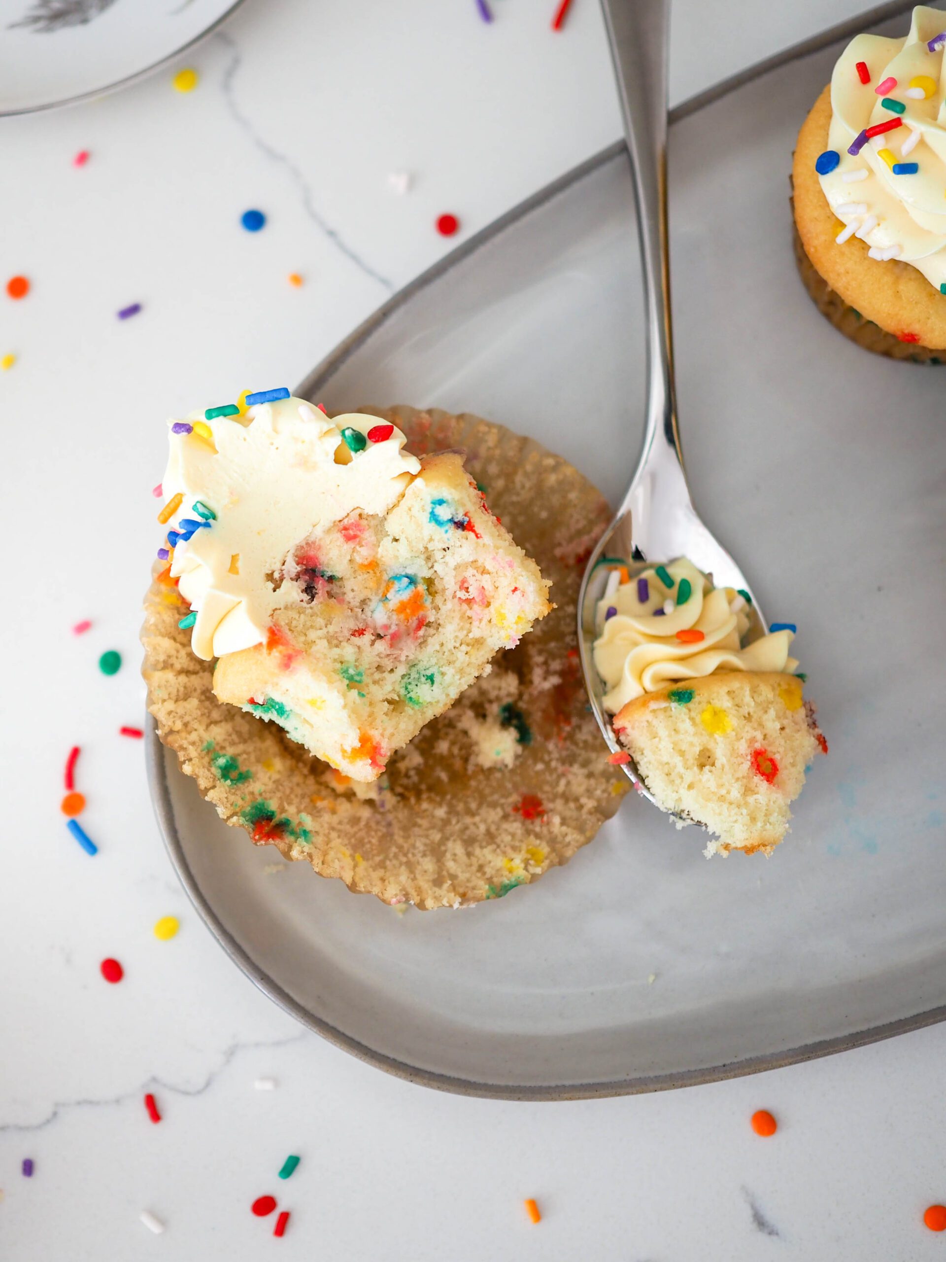 A spoon has removed a bite of confetti cupcake, with the spoon remaining nearby it on a plate.