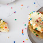 A confetti cupcake lays on its side with a spoonful taken out. Sprinkles surround it on a white quartz counter.
