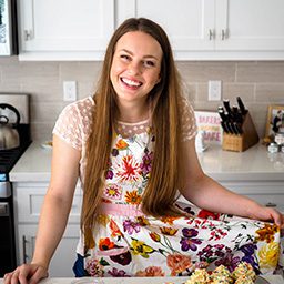 Alyssa, a white woman with brown hair, smiles while holding up a corner of her floral apron.