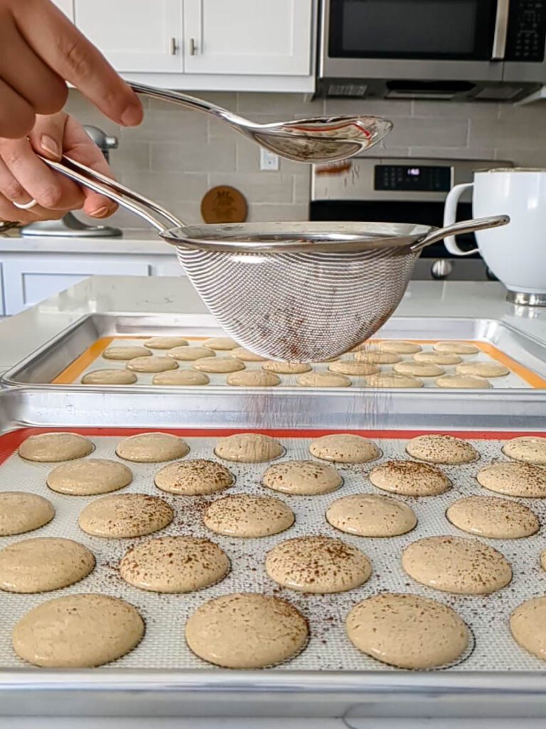 A fine mesh strainer dusts cocoa over the tops of one tray of macaron shells.
