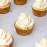 One confetti cupcake with a swirl of off-white buttercream on top is in focus. Other cupcakes are around it.