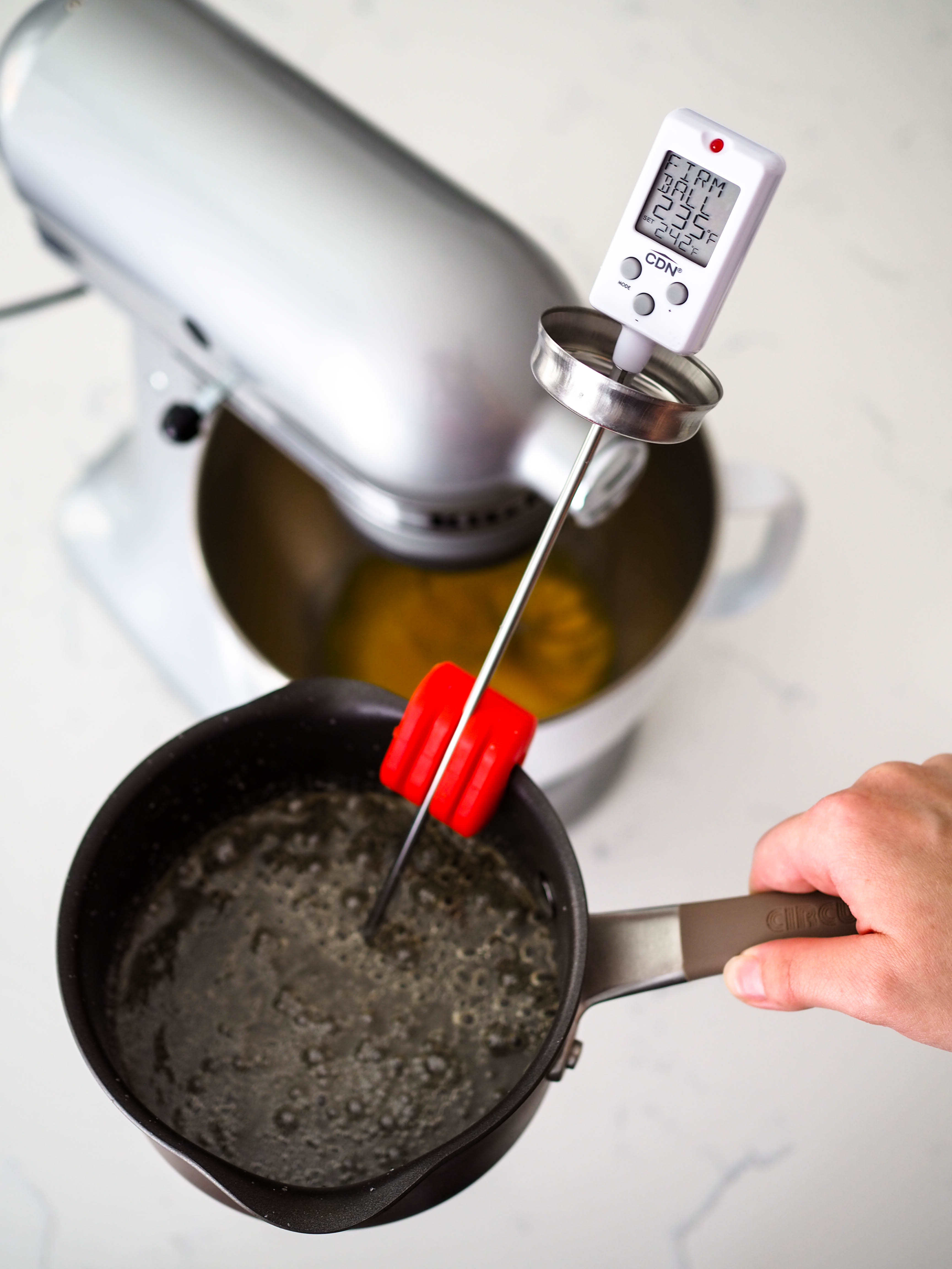 A candy thermometer shows the temperature of the sugar syrup in front of a stand mixer.