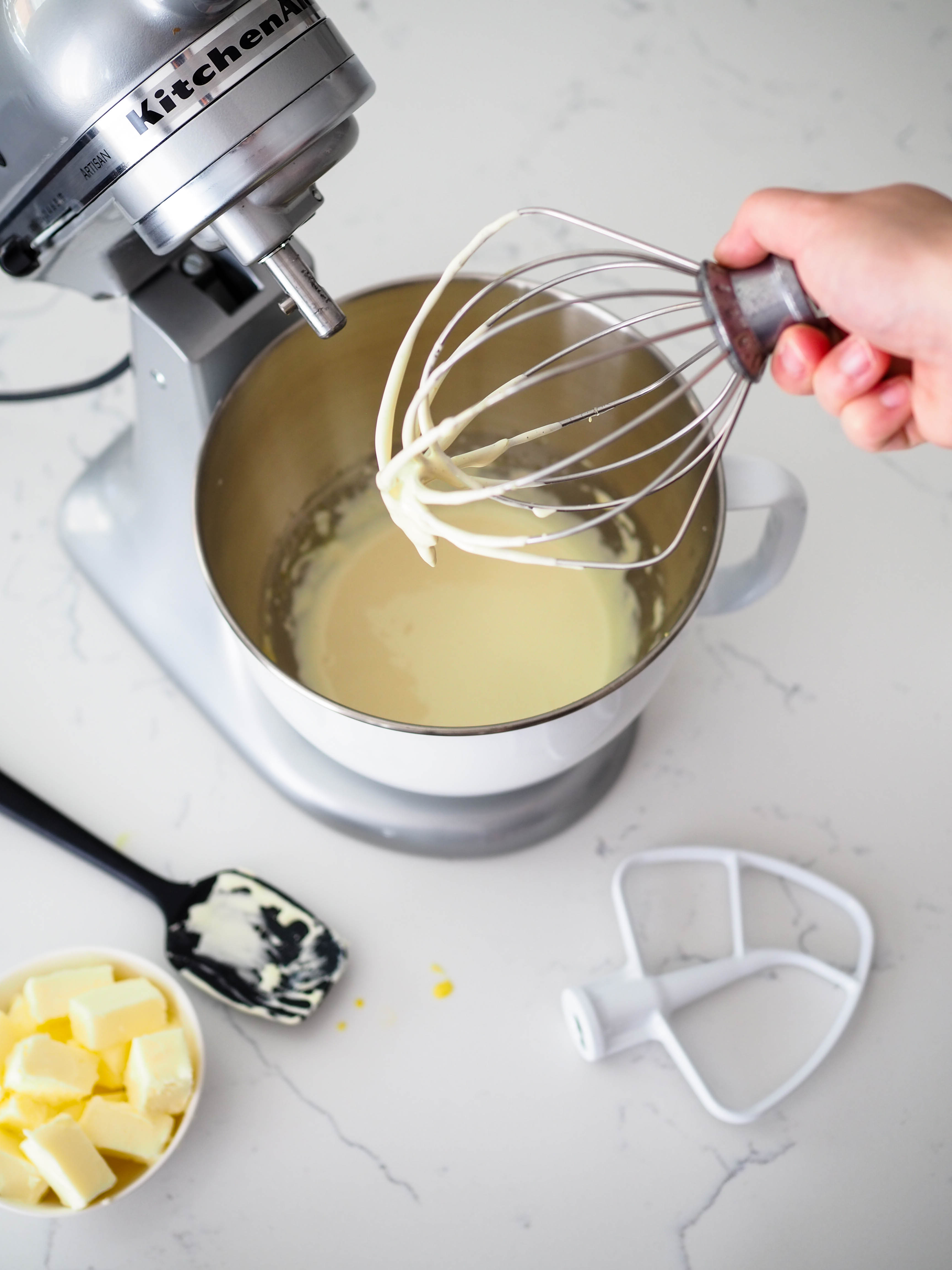 A whisk with custard filling the bottom in front of a stand mixer.