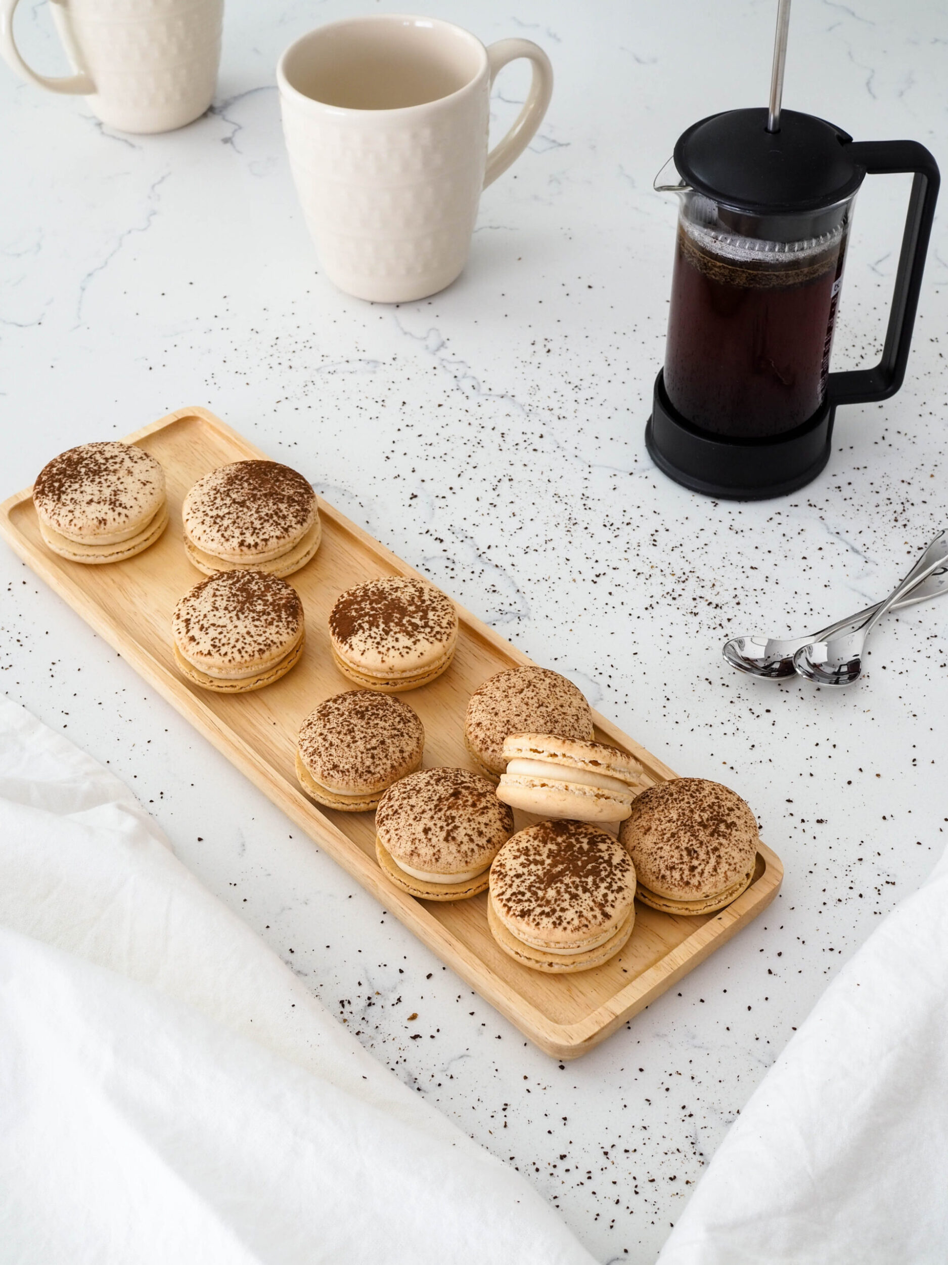 Tiramisu macarons on a wooden platter, with a French press filled with coffee nearby.